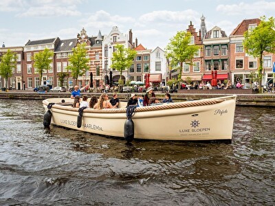 Een luxe sloep vol met passagiers vaart door de grachten van Haarlem, met historische gebouwen en terrasjes op de achtergrond.